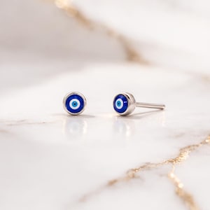 Könnte beinhalten: Ein Paar silberne Ohrstecker mit einem blau-weißen Evil-Eye-Design. Die Ohrringe haben eine runde Form mit einem kleinen Stift zum Durchstechen. Das Evil-Eye-Symbol ist ein Schutz-Talisman.