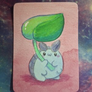 May include: A watercolor illustration of a cute, light blue creature with large ears, holding a large green leaf. The background is a soft pink, and the image is on a rectangular card.