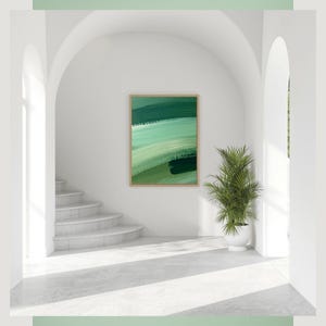 Puede incluir: Impresión artística abstracta enmarcada con pinceladas verdes. La obra de arte cuelga de una pared blanca en una habitación luminosa con una escalera y una planta en maceta. El marco es de color madera clara.