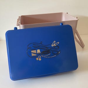 May include: A rectangular sewing box with a light pink base and a detachable blue lid. The lid features a gold-coloured graphic illustration of sewing supplies, including scissors, a spool of thread, a thimble, a measuring tape, and buttons.