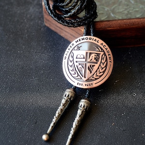 May include: A bolo tie with a circular, copper-colored pendant featuring the words "WALKER MEMORIAL ACADEMY" and "EST. 1947". The pendant has a shield design with a cross, book, and eagle. The tie has a black braided cord and silver-colored tips.