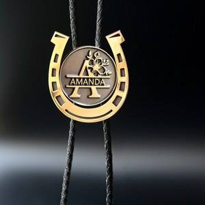 May include: A gold horseshoe bolo tie with the letter A and the name Amanda engraved on the horseshoe. The bolo tie is on a black background.