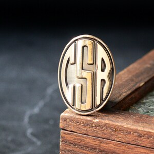 May include: A gold-colored, oval-shaped pin with the letters "CSR" in a stylized, overlapping design. The pin is set against a dark background and rests on a wooden surface, showcasing its metallic sheen and intricate details.