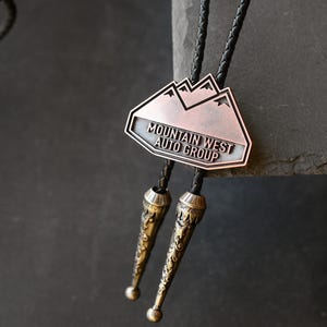 May include: A bolo tie with a copper-coloured metal pendant featuring a mountain design and the text "MOUNTAIN WEST AUTO GROUP." The tie has a black braided cord and decorative metal tips.