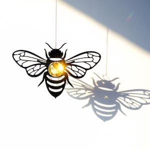 May include: A black metal bee silhouette with a crystal in the centre hangs from a silver chain. The bee casts a shadow on a white wall.