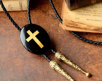 Op de afbeelding: Een zwarte bolo stropdas met een gouden kruisontwerp. De ovale hanger is verbonden met een gevlochten zwart koord met decoratieve goudkleurige uiteinden. De achtergrond toont open boeken.