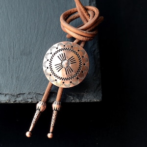 May include: A copper bolo tie with a circular pendant featuring an etched sunburst design. The pendant is connected to a brown leather cord, with decorative metal tips. The cord is knotted at the top.