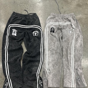 Może przedstawiać: Dwie pary spodni dresowych w kolorze antracytowym i szarym. Każda para ma białe paski po bokach, zamki błyskawiczne przy kostkach i haftowane motywy religijne. Tekst "HOLY" i "HOLY SPIRIT" jest widoczny na parze w kolorze antracytowym.