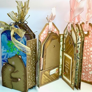 May include: Handmade tag albums with decorative covers. One album features a bear design with a blue and white background, and gold ribbon. Other albums have floral and wood grain patterns, with pink ribbon ties. One album has the text "It's the little things in life."