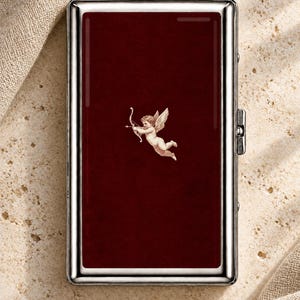 May include: A rectangular metal cigarette case with a polished silver frame and a deep burgundy center panel. The center features a small, detailed illustration of a winged cherub holding a bow and arrow, centered on the dark red surface.