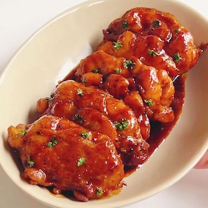 Puede incluir: Un plato de muslos de pollo glaseados en un plato ovalado beige. El pollo está cubierto con una salsa brillante de color marrón rojizo y adornado con hierbas verdes frescas. El plato se presenta sobre una superficie blanca.