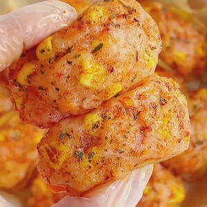May include: Close-up of seasoned chicken breasts, held in a gloved hand. The chicken is coated in a blend of spices, with visible herbs and a vibrant orange hue. The texture appears moist and flavorful, suggesting a delicious meal.