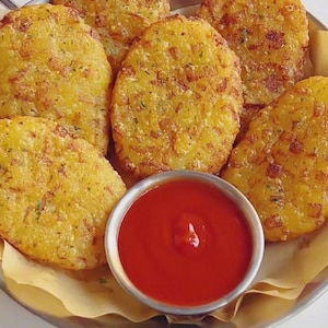 May include: A plate of golden potato cakes, served with a small bowl of red ketchup. The potato cakes are round and crispy, with visible herbs and spices. The food is presented on a metal plate, a classic comfort food.