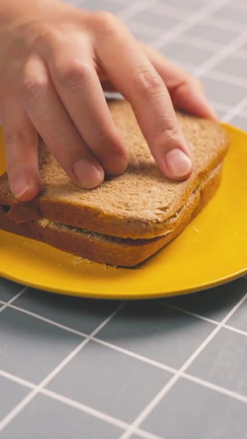 May include: A sandwich on a bright yellow plate. The sandwich is made with whole wheat bread and appears to have a filling. A hand is reaching for the sandwich.
