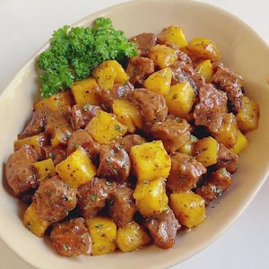 Puede incluir: Primer plano de un plato ovalado beige lleno de una comida abundante. El plato contiene trozos de carne cocida y patatas de color amarillo dorado, adornado con perejil verde fresco. La comida parece recién preparada.