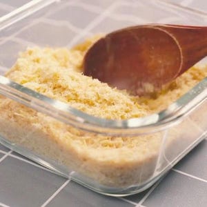 May include: A clear glass container filled with a light yellow, shredded food product. A wooden spoon rests inside the container, partially submerged in the food. The container sits on a gray and white checkered surface.