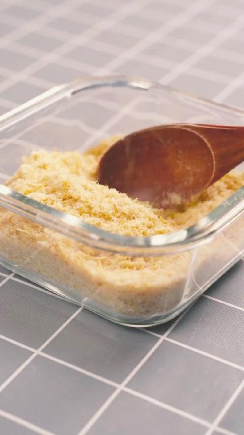 May include: A clear glass container filled with a light yellow, shredded food product. A wooden spoon rests inside the container, partially submerged in the food. The container sits on a gray and white checkered surface.