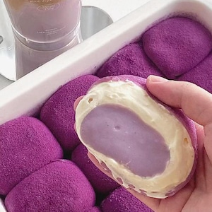 Puede incluir: Una bandeja de pasteles morados tipo mochi con un relleno cremoso, uno cortado para revelar el interior. Un vaso de café helado con el texto "Cafe de La vie" está en el fondo. Los pasteles son de un color morado vibrante.