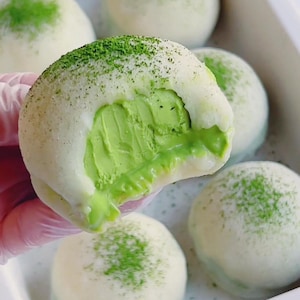Puede incluir: Primer plano de un helado mochi de matcha con un mordisco, que revela el relleno de helado verde. El mochi es blanco con polvo de matcha verde espolvoreado por encima. Hay otras bolas de mochi en el fondo.