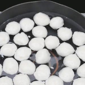 Può includere: Primo piano di una padella nera piena di acqua limpida e numerosi alimenti bianchi e rotondi. Gli alimenti sembrano essere in cottura o in ammollo nell'acqua. Lo sfondo è di colore nero pieno.