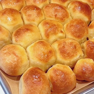 Puede incluir: Una bandeja de panecillos dorados recién horneados. Los panecillos están colocados sobre una hoja de papel de pergamino. Tienen un aspecto brillante y ligeramente caramelizado, lo que sugiere que son suaves y mantecosos.