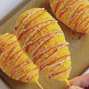 Könnte beinhalten: Drei goldgelbe Corn Dogs auf einem Backblech, beträufelt mit roten und weißen Saucen. Die Corn Dogs sind auf Holzstäbchen und haben eine knusprige, strukturierte Beschichtung. Mit grünen Kräutern bestreut.