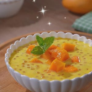 May include: A white bowl of mango sago dessert, a creamy yellow pudding with small tapioca pearls and diced mango pieces. Garnished with mint leaves, the dessert sits on a wooden tray.