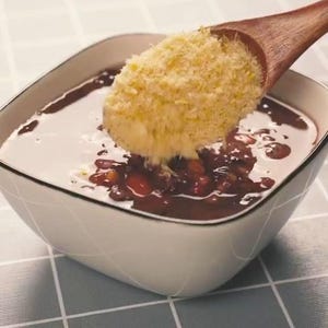 May include: A square white bowl with a dark red liquid and beans, topped with a yellow crumble. A wooden spoon is pouring the crumble into the bowl. The bowl sits on a gray and white checkered surface.