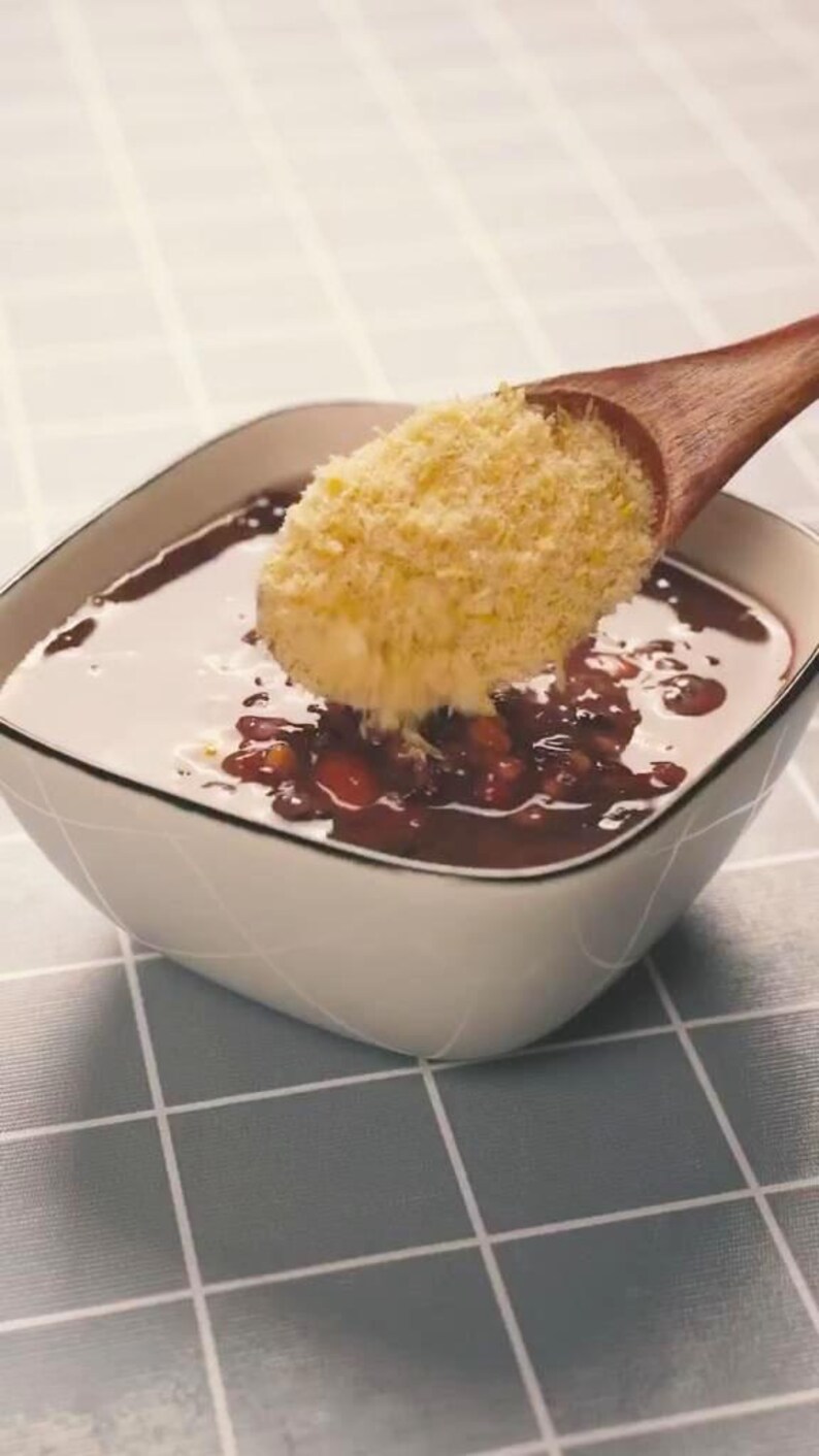 May include: A square white bowl with a dark red liquid and beans, topped with a yellow crumble. A wooden spoon is pouring the crumble into the bowl. The bowl sits on a gray and white checkered surface.