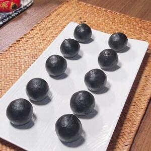 Puede incluir: Un plato rectangular blanco contiene una colección de alimentos oscuros y redondos. Parecen ser un tipo de postre o aperitivo, con un acabado liso y mate. El plato está colocado sobre un mantel individual tejido, lo que sugiere un ambiente informal.