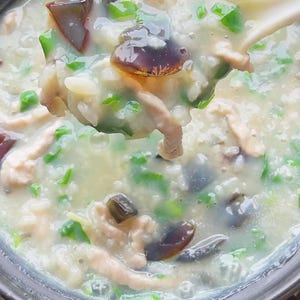 May include: A close-up of a bowl of congee, a rice porridge, with visible ingredients. The porridge contains pieces of meat, dark-coloured eggs, and green herbs. A spoonful of the congee is being lifted from the bowl.