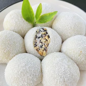 May include: A plate of mochi, a Japanese rice cake, covered in shredded coconut. One mochi is cut open, showing a filling of black sesame seeds and nuts. Green leaves are placed on top of the mochi.