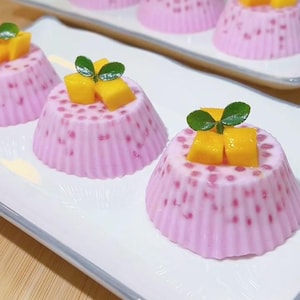 May include: A close-up of several pink desserts with a mango topping and green leaves, arranged on a white rectangular plate. The desserts have a textured, dotted appearance and are set on a wooden surface.