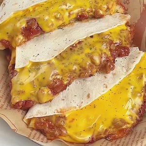 May include: Overhead shot of three quesadillas with melted yellow cheese and a red filling, served on a paper-lined plate. The tortillas are folded in half, showcasing the cheesy filling.