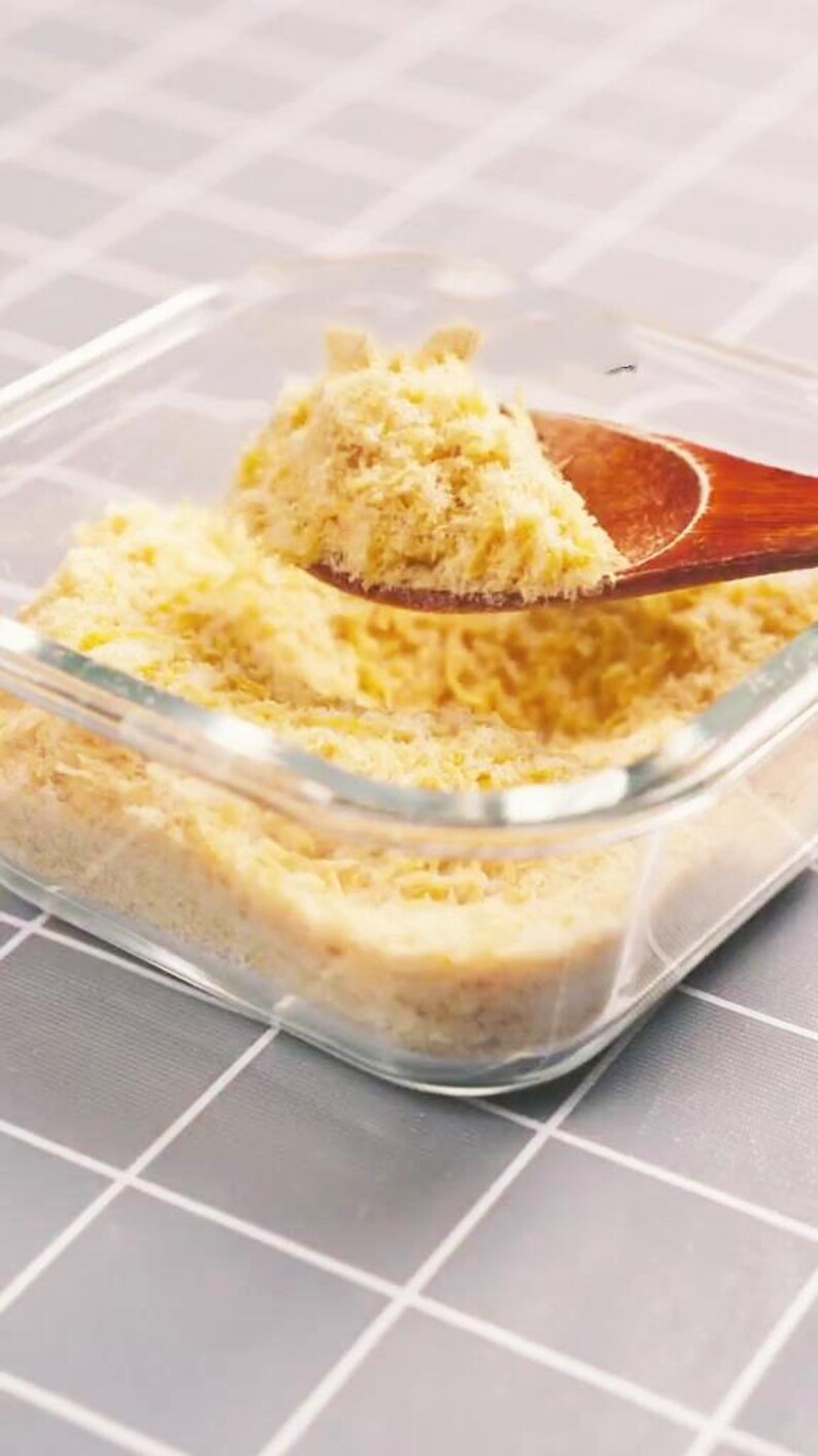 May include: A clear glass dish filled with yellow breadcrumbs. A wooden spoon scoops up a portion of the breadcrumbs. The dish sits on a gray and white checkered surface. The breadcrumbs appear to be freshly made.