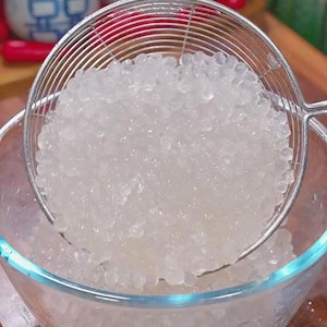 May include: A clear glass bowl filled with translucent tapioca pearls, with a metal mesh strainer scooping up a portion of the pearls. The bowl sits on a wooden surface, with decorative items in the background.