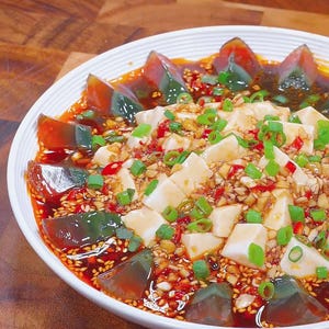 May include: A white bowl of food with dark, triangular eggs, white tofu cubes, and a red sauce. Garnished with green onions and sesame seeds, the dish sits on a wooden surface. A colourful meal.