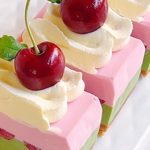 Op de afbeelding: Close-up van gelaagde dessertschijfjes met een groene basis, een roze middenlaag en witte slagroomtopping. Elke schijf is gegarneerd met een verse, rode kers en een groen blad.