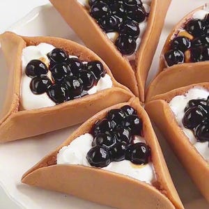 Puede incluir: Un plato blanco con seis pasteles triangulares, cada uno relleno de crema blanca y cubierto con perlas de tapioca oscuras y brillantes. Los pasteles tienen un exterior marrón claro y ligeramente texturizado.