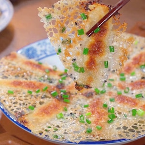 Puede incluir: Primer plano de una empanadilla crujiente, dorada y marrón, con una textura delicada y calada, adornada con cebollino verde fresco y semillas de sésamo negro. La empanadilla está sujeta por palillos de madera, revelando su sabroso relleno. Está en un cuenco azul y blanco.