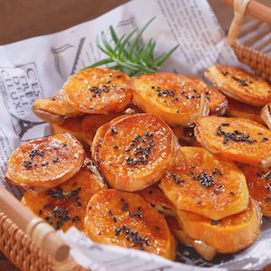 May include: A close-up shot of a basket filled with glazed sweet potato slices, sprinkled with black sesame seeds. The sweet potatoes are arranged on a piece of paper with text. A sprig of rosemary is placed on top.
