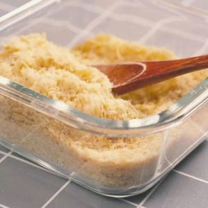 May include: A clear glass container filled with a light yellow, granular substance. A wooden spoon rests inside the container, partially submerged in the substance. The container sits on a gray and white checkered surface.