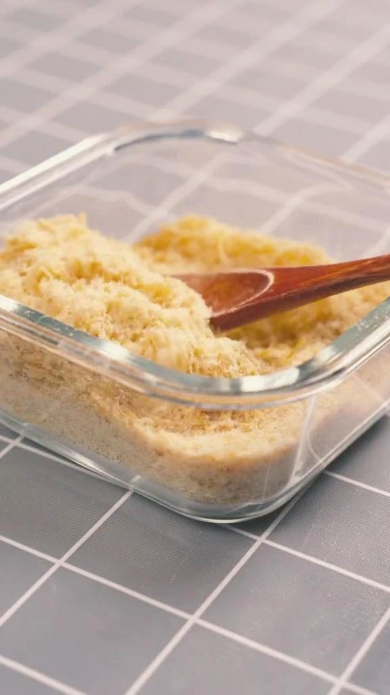 May include: A clear glass container filled with a light yellow, granular substance. A wooden spoon rests inside the container, partially submerged in the substance. The container sits on a gray and white checkered surface.