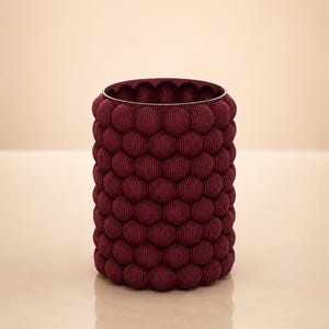 May include: A cylindrical burgundy-colored container with a textured, bubble-like pattern. The top rim is a lighter shade of burgundy. The container is likely used for storage or decoration.