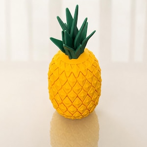 May include: A yellow pineapple-shaped decorative object with a textured surface. The pineapple has green leaves at the top. The object is sitting on a reflective surface.