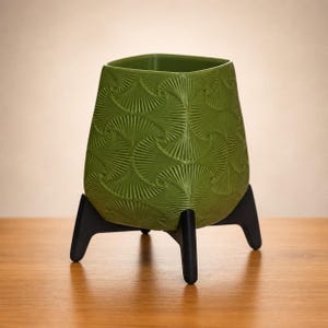 May include: A green ceramic planter with a textured, fan-like pattern. The planter sits on a black, three-legged stand. The square-shaped top of the planter contrasts with the rounded base, creating a modern aesthetic.