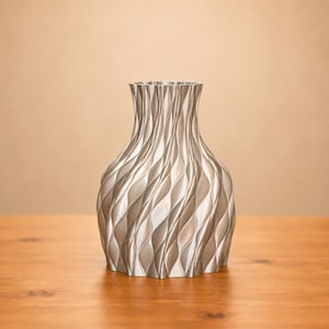 May include: A silver-colored vase with a textured, wave-like pattern. The vase has a wide base that tapers to a narrower neck and flared rim. The vase is approximately 8 inches tall and sits on a wooden surface.