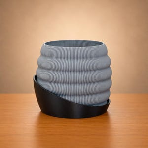 May include: A gray, ribbed planter with a black base. The planter has a cylindrical shape with horizontal ridges. The black base is a curved, dish-like shape. The planter is sitting on a wooden surface.