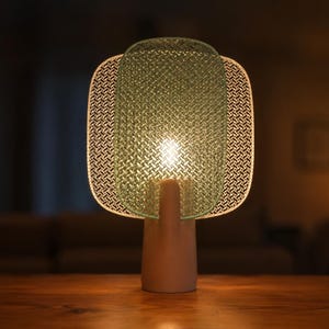 May include: A modern table lamp with a cylindrical base and a unique shade. The shade features a green, woven center and a patterned, off-white border. The lamp is illuminated, casting a warm glow.