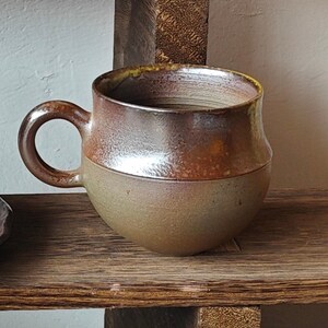 May include: A handmade ceramic mug with a rounded base and a sturdy handle. The mug features a two-tone design, with a light brown lower half and a darker, metallic-looking upper half. The rim has a yellow-green accent.